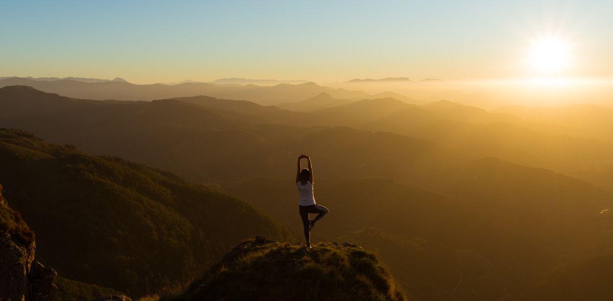 Woman on top of mountain practising the yoga asana the tree with view over the summits during sunset